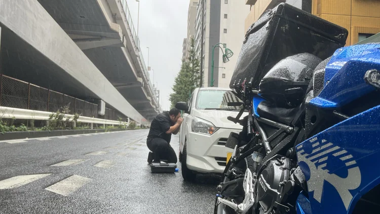 雨の路上で車の鍵穴に向き合う隊員と、手前に停めた青いバイク