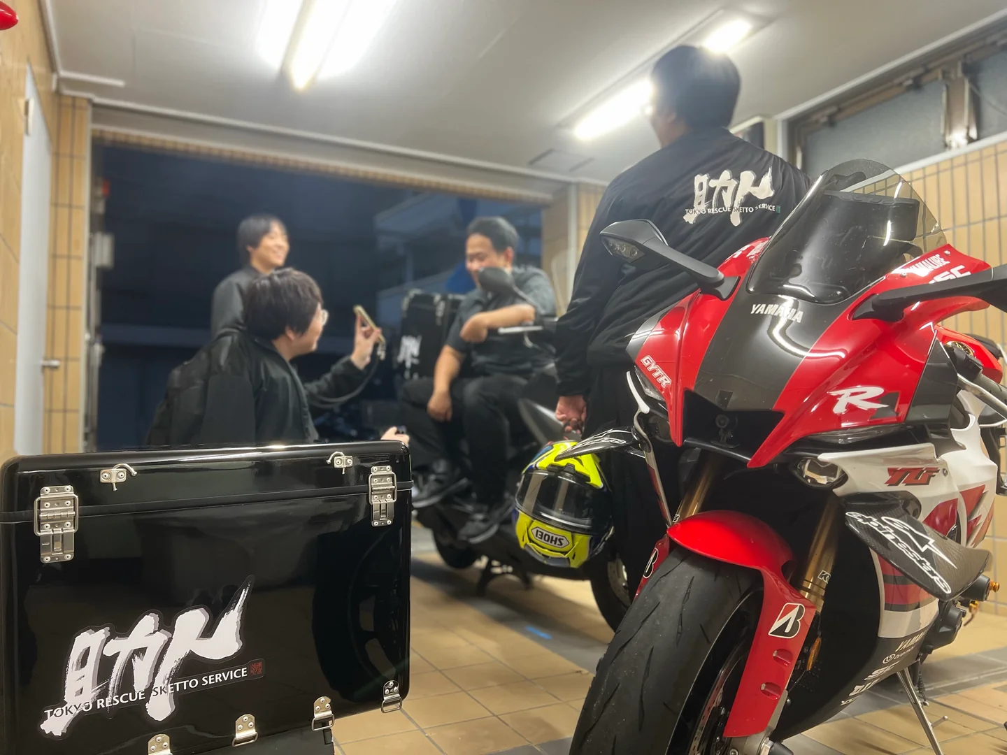 救援用バイクや工具箱のある工房で、助人の隊員たちが話している様子